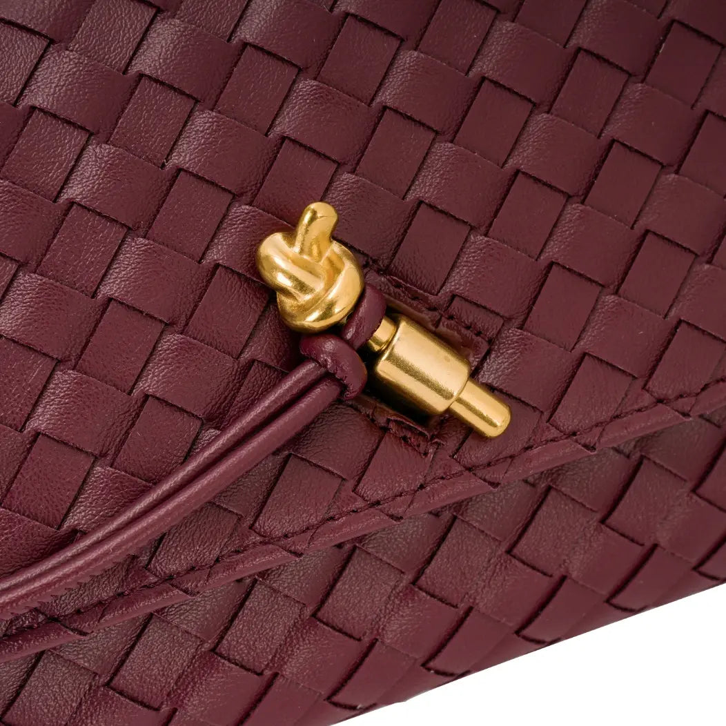 Detalhe aproximado do adorno metálico dourado em formato de nó e do acabamento trançado da aba frontal da bolsa clutch Diana vinho. Preto Bordô