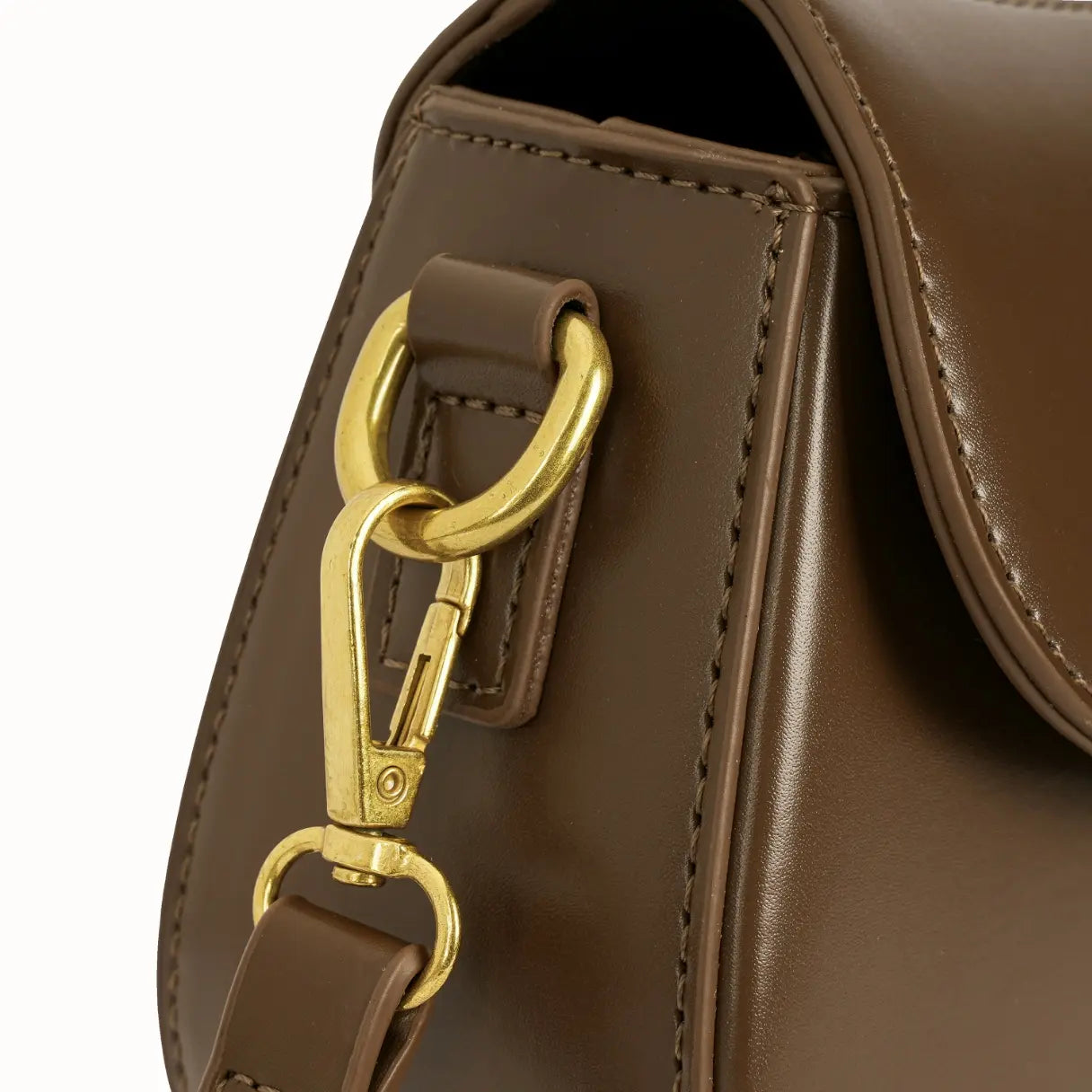 Detalhe em zoom do mosquetão metálico dourado prendendo a alça longa transversal na argola lateral revestida em couro da bolsa. Preto Caramelo Off White Marrom