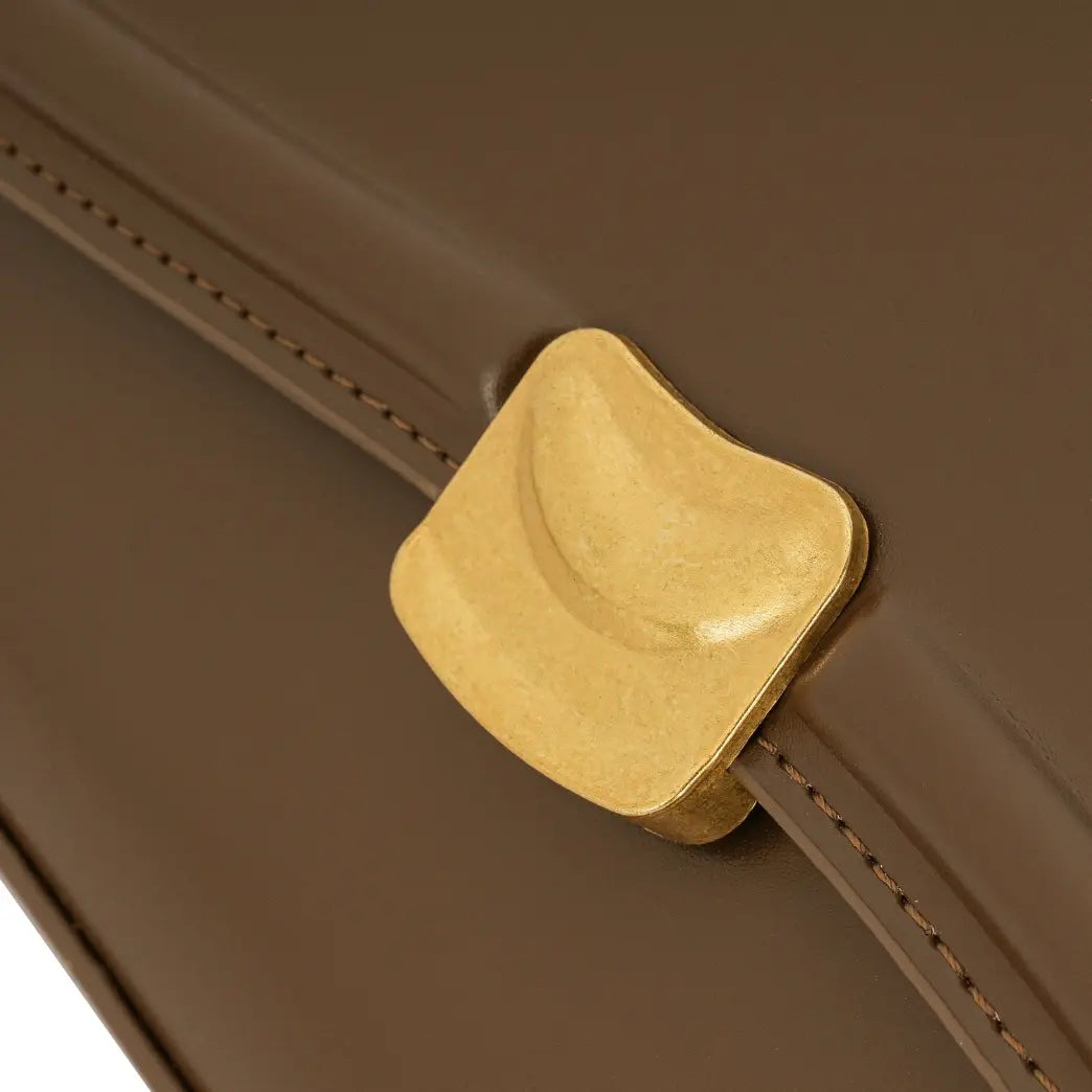 Detalhe em zoom do fecho magnético de metal dourado escovado na aba frontal da Bolsa Baguete Sophia marrom. Preto Caramelo Off White Marrom