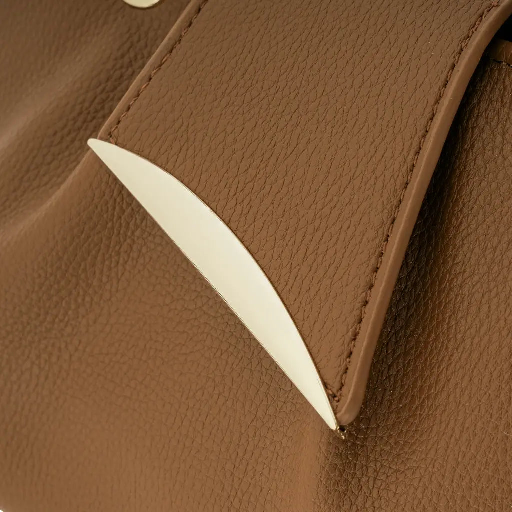 Detalhe em zoom do fecho metálico dourado liso com design curvo e minimalista na aba frontal da Bolsa Mini Tote Luna marrom. Preto Caramelo Off White