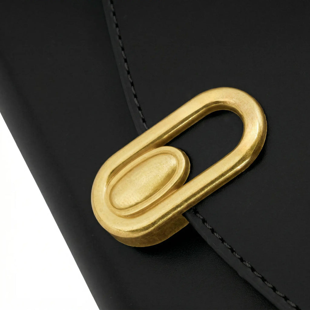 Close no fecho de metal dourado com design oval minimalista na aba frontal da Bolsa Baguete Vivianne preta. Preto [cor:Preto]