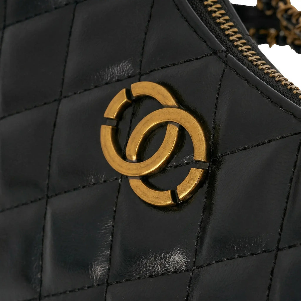 Detalhe do emblema metálico dourado em formato de elos entrelaçados sobre o couro matelassê da Bolsa Amelia. Preto [cor:Preto]