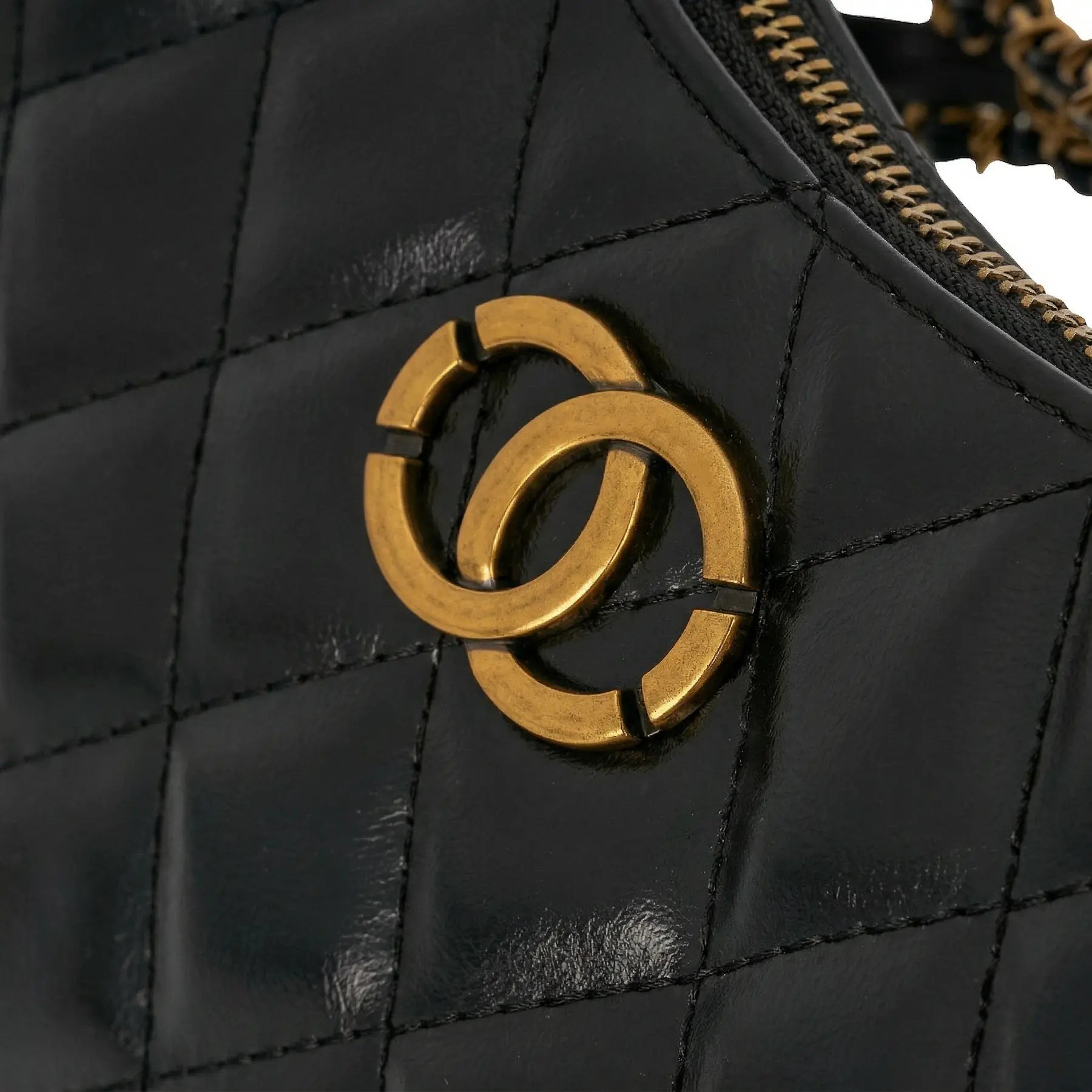 Detalhe do emblema metálico dourado em formato de elos entrelaçados sobre o couro matelassê da Bolsa Amelia. Preto [cor:Preto]